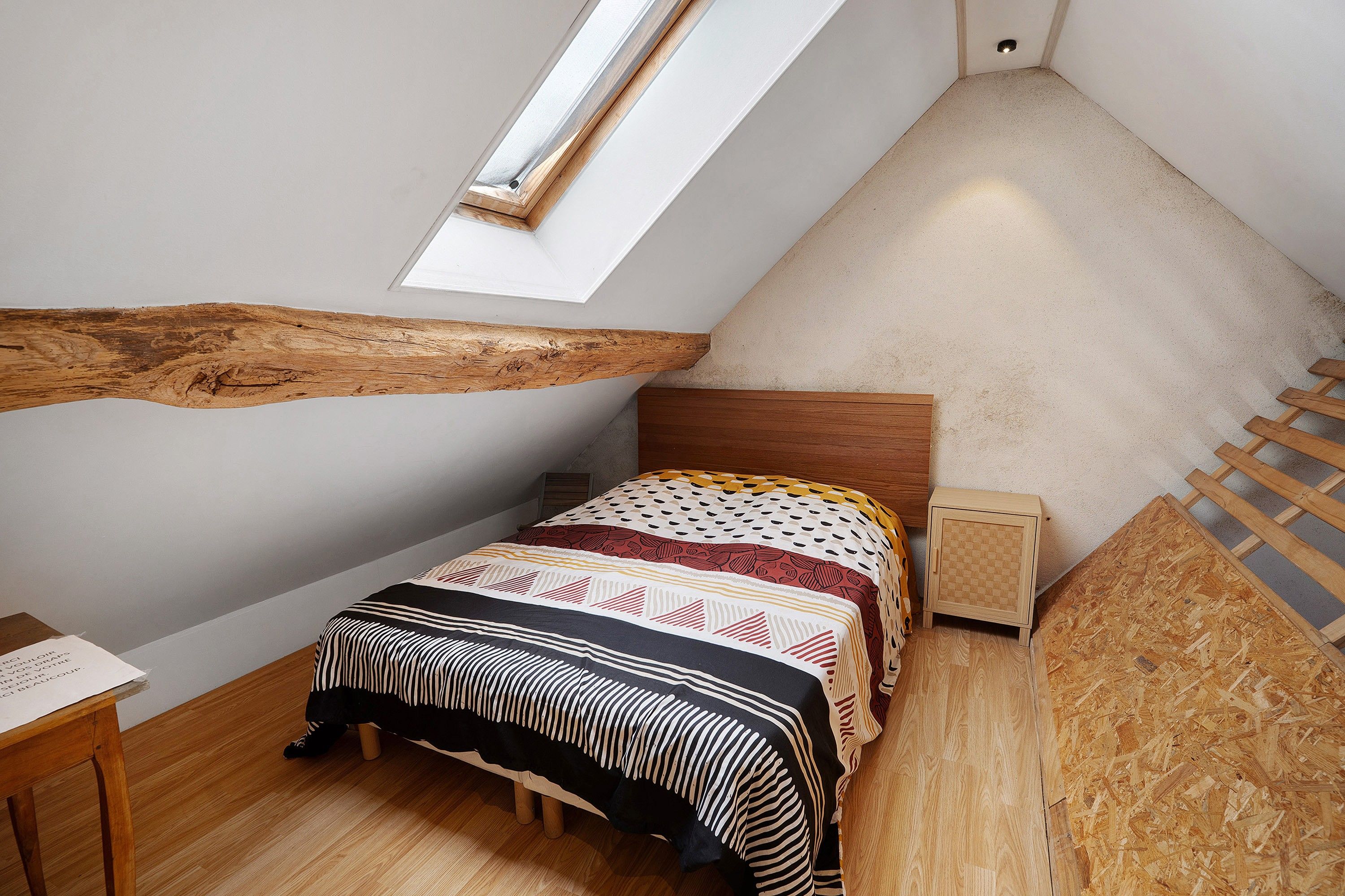 Een kleine, schuine kamer met een enkel bed, een houten hoofdbord, een klein nachtkastje en een dakraam.