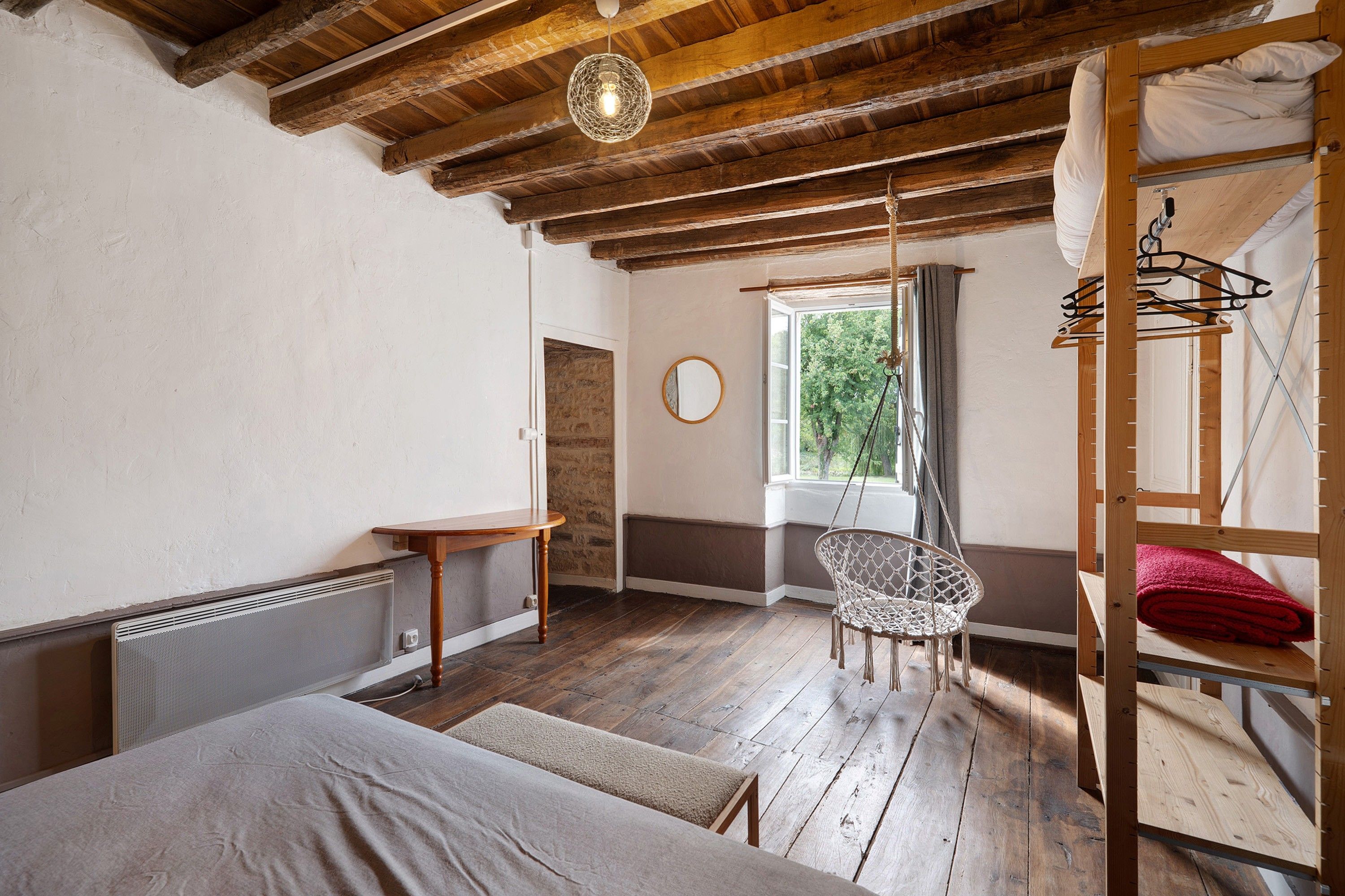 Une pièce rustique avec des poutres apparentes, un lit, une chaise suspendue, une fenêtre donnant sur les arbres et un radiateur.
