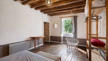 Une pièce rustique avec des poutres apparentes, un lit, une chaise suspendue, une fenêtre donnant sur les arbres et un radiateur.