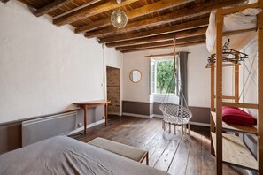 Une pièce rustique avec des poutres apparentes, un lit, une chaise suspendue, une fenêtre donnant sur les arbres et un radiateur.