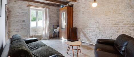 A cozy living room with a stone wall, a wooden ceiling, a black leather sofa, a wooden chair, and a floor lamp.