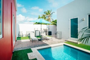 A modern outdoor pool area with a blue pool, lounge chairs, a barbecue grill, and a small garden with artificial grass.