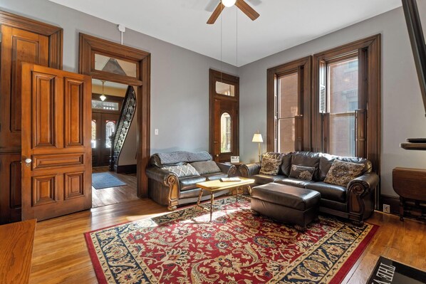 A spacious living room with a large wooden door, a leather sofa, a coffee table, and a red patterned rug.