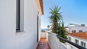 Un balcón estrecho con suelo de ladrillo rojo, paredes blancas y vistas al mar.