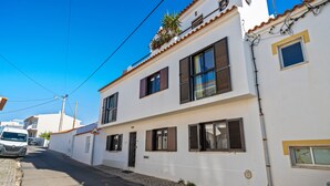 Un edificio de dos pisos con fachada blanca, varias ventanas con contraventanas y una calle adoquinada.