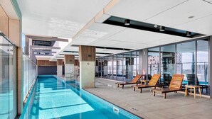 An indoor swimming pool with clear water, wooden loungers, and glass walls offering views of the city. - Luxe on Franklin | Car Park, Lap Pool, Sauna, BBQ (Adelaide)