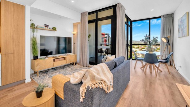 A modern living room with a large flat-screen TV, a sofa, a dining area with a table and chairs, and a balcony with a view of the city. - Luxe on Franklin | Car Park, Lap Pool, Sauna, BBQ (Adelaide)