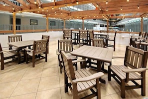A covered outdoor dining area with wooden tables and chairs, a tiled floor, and a view of a building in the background.