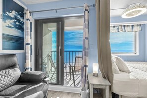 A room with a bed, a chair, a table with a lamp, a balcony with a view of the ocean, and a painting of the ocean on the wall.