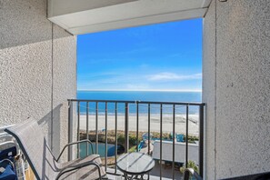 A balcony with a view of the beach, a pool, and outdoor furniture.