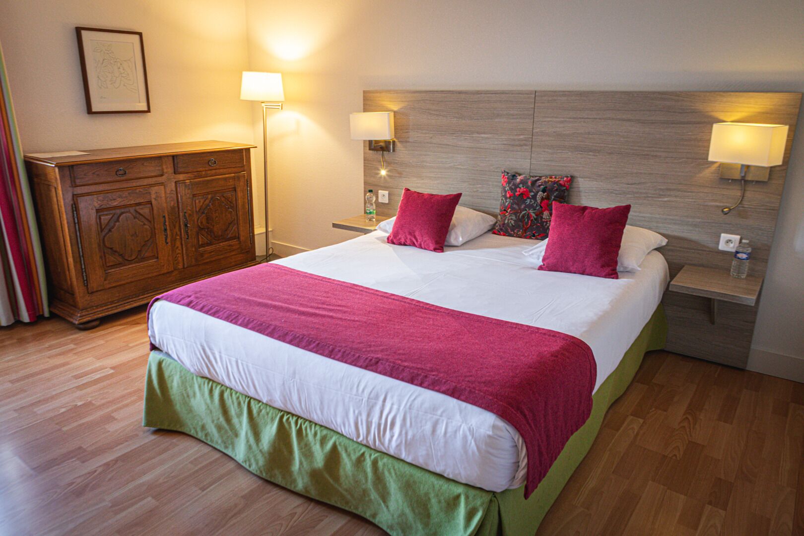 Une chambre d’hôtel avec un grand lit, une tête de lit en bois, des lampes de chevet, une commode et un tableau encadré au mur.