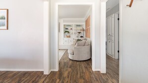 Un pasillo con piso de madera, un sillón beige y un mueble blanco con estantes y armarios.