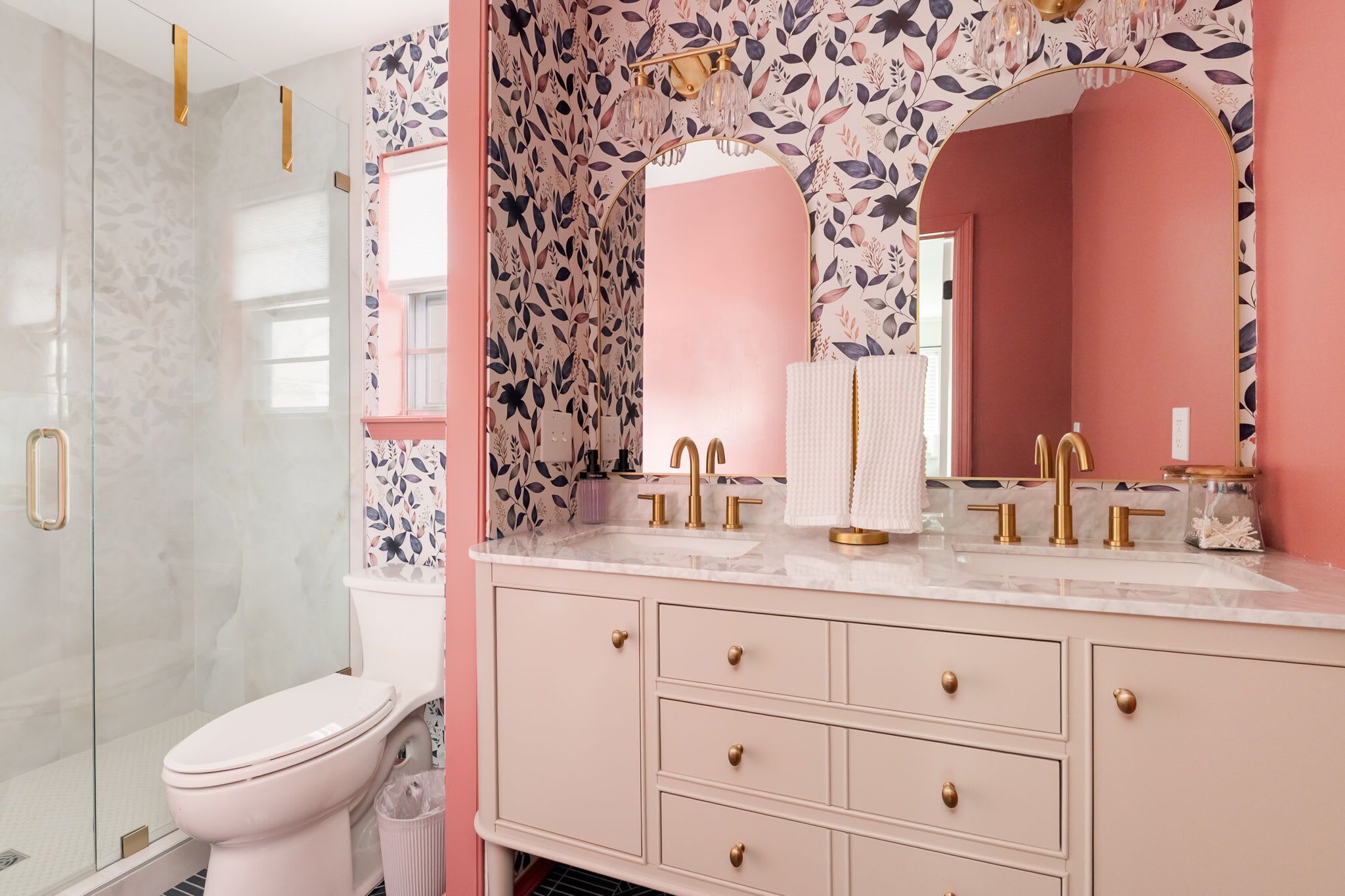 Une salle de bain aux couleurs rose et blanc, dotée d’une douche en verre, de toilettes, d’un meuble-lavabo avec des robinets dorés et d’un miroir orné de motifs floraux.