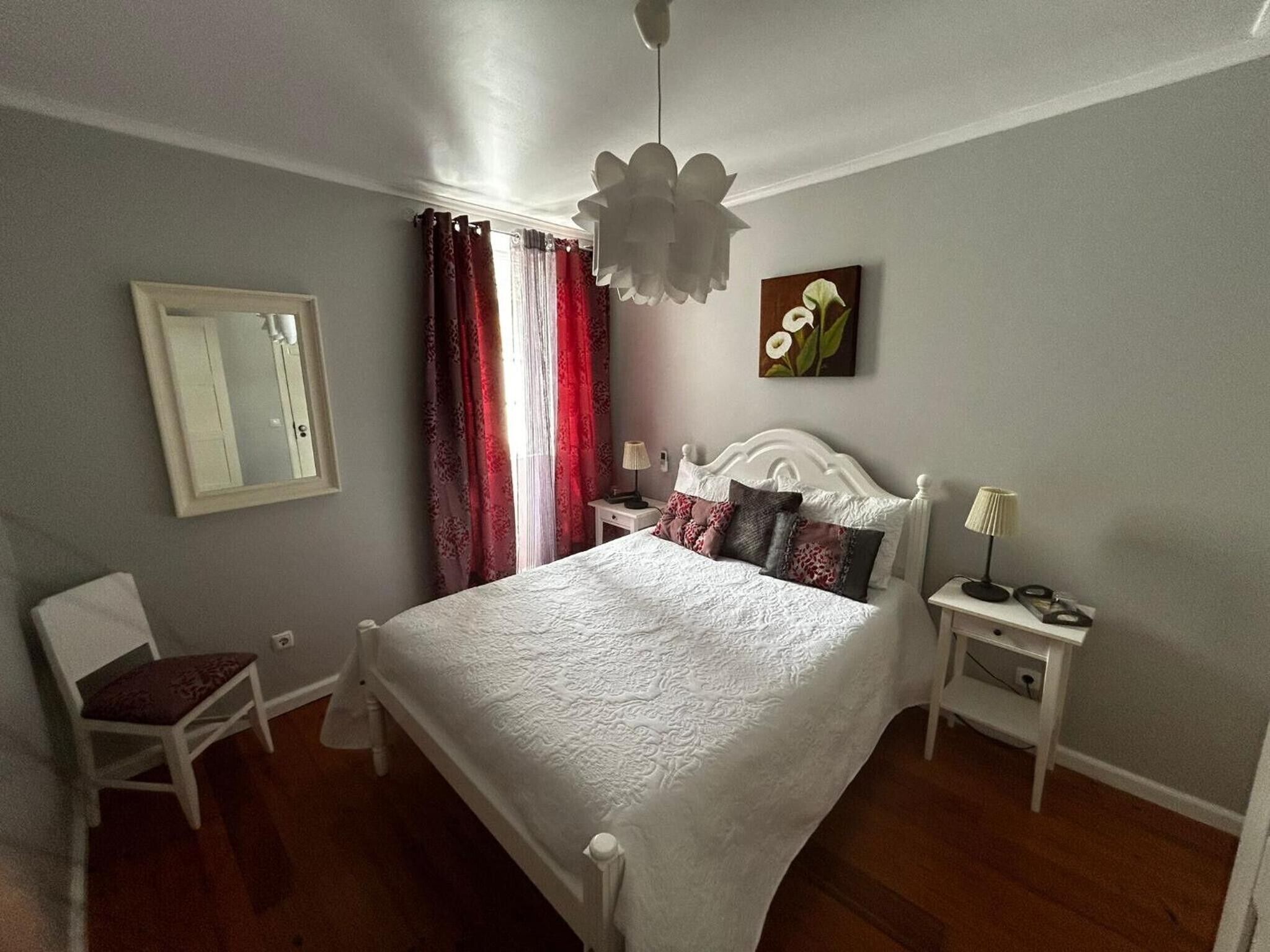 Un dormitori amb llit blanc, una cadira, una tauleta de nit amb làmpada, un mirall i un quadre a la paret.