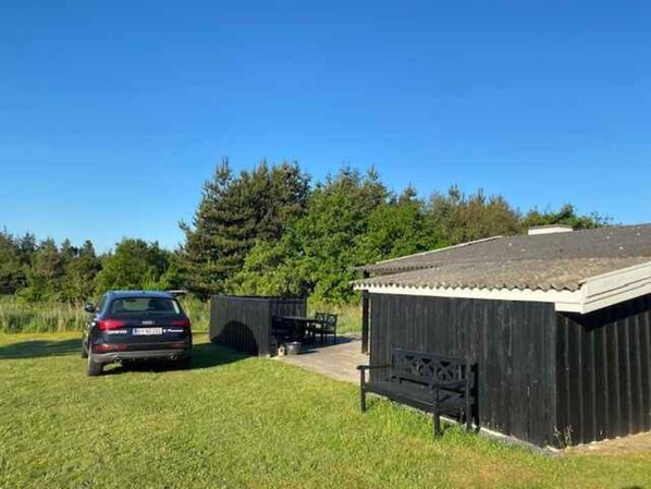 A small, dark wooden structure with a sloped roof, a bench outside, and a car parked nearby.