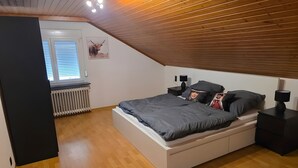 Une chambre avec un plafond en pente, un lit avec une literie grise, deux tables de chevet avec des lampes, un radiateur et une fenêtre avec des stores.