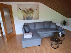 A modern living room with a grey sectional sofa, a wooden floor, a door, a wall-mounted bull painting, a small round table with a keyboard, and two potted plants.