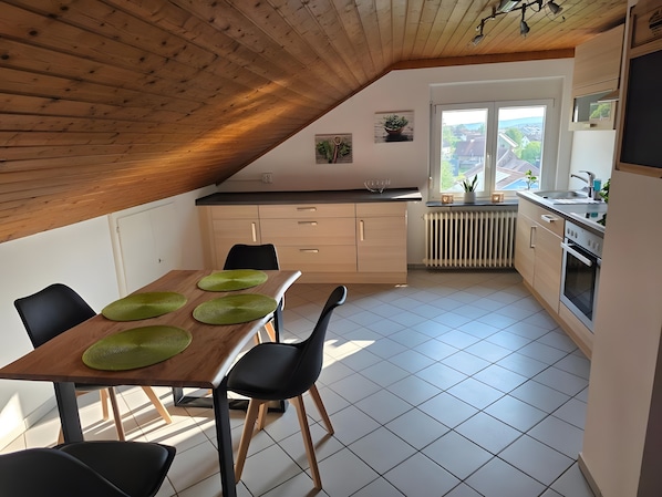 A modern kitchen with a wooden ceiling, a dining table with green placemats, and a view of the outside through a window.