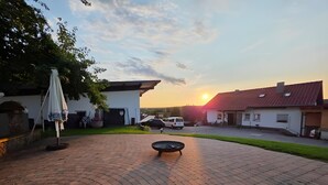 Une cour dallée avec un foyer circulaire, un parasol blanc et un fourgon blanc garé au fond.