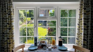 A breakfast table with a view of a garden through large windows, a coffee maker, and a basket of bread.