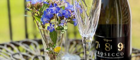 Une bouteille de vin rosé, deux verres à vin transparents et un petit vase avec des fleurs bleues, le tout posé sur une table en métal à l’extérieur.