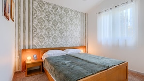 Ein Schlafzimmer mit einem hölzernen Bett, einem Nachttisch, einer Lampe, einer Wand mit Musterung sowie einem Fenster mit Vorhängen.