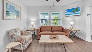A living room with a brown sofa, a patterned armchair, a wooden coffee table, and a ceiling fan. There are two windows with blinds, a framed picture on the wall, and a TV mounted above the sofa. - ☀️ Pet Friendly - Entire Property - The Bungalows ☀️ (Treasure Island)