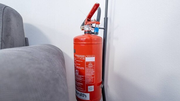 A red fire extinguisher mounted on a wall next to a gray couch. - [Modern] 2BR/2BA • Metro Access • 12min Duomo (Milan)