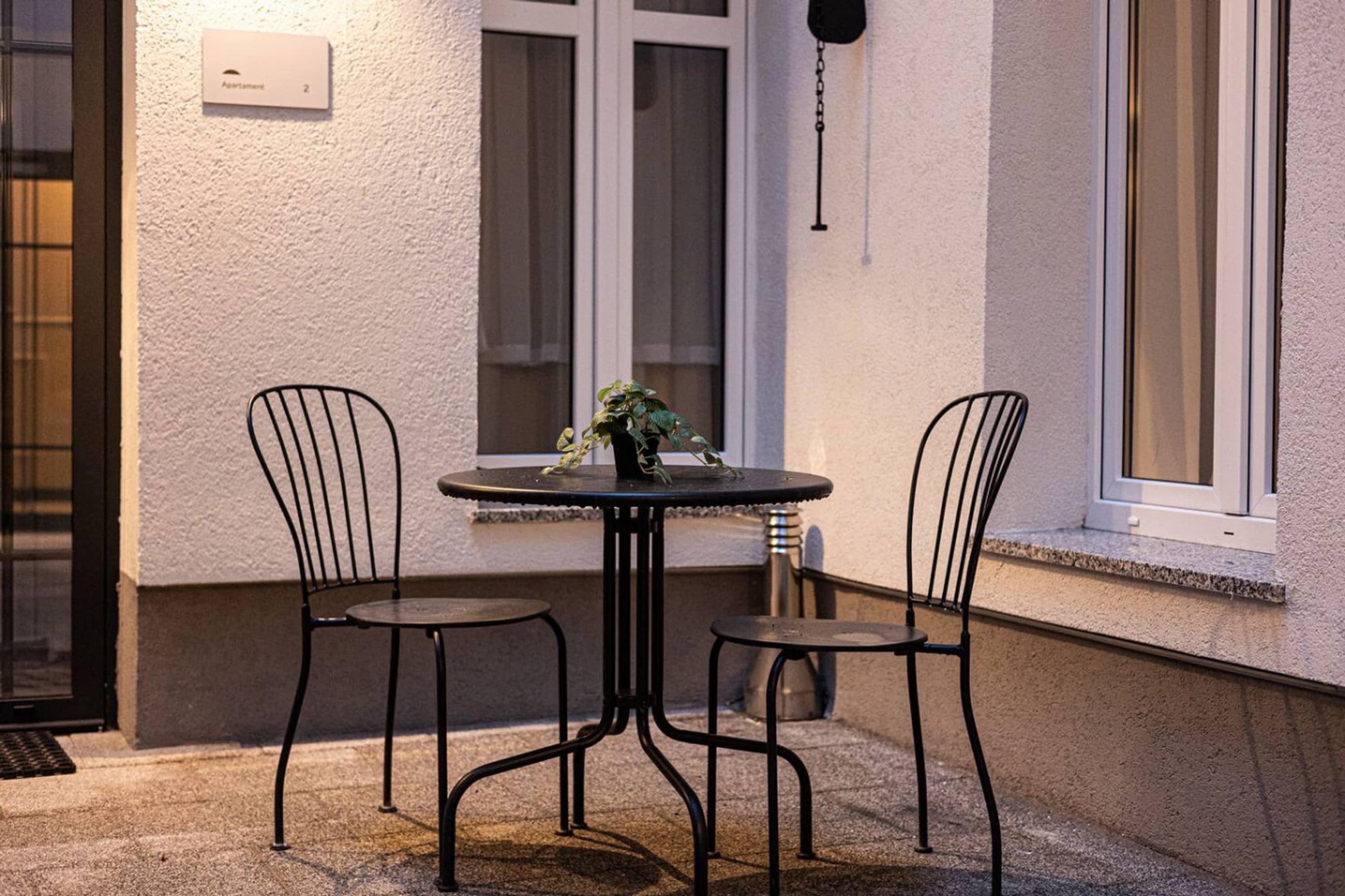 Uma pequena área externa com mesa redonda e duas cadeiras, um vaso de planta na mesa e uma janela com persianas.