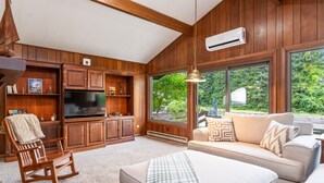 Una sala de estar acogedora con paneles de madera, un ventanal con vistas a la vegetación, un sofá, un sillón mecedora y un televisor.