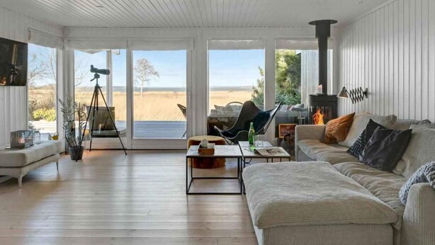 A spacious living room with a large window overlooking a beach, a fireplace, and a telescope on a tripod. - 5 Star Holiday Home in Hillerod (Hillerod)