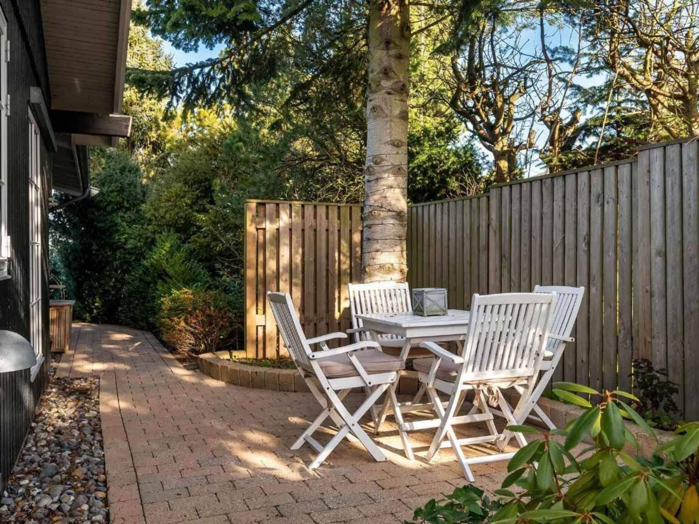 Eine Terrasse mit Holztisch und weißen Stühlen, umgeben von einem Holenzaun und Bäumen.