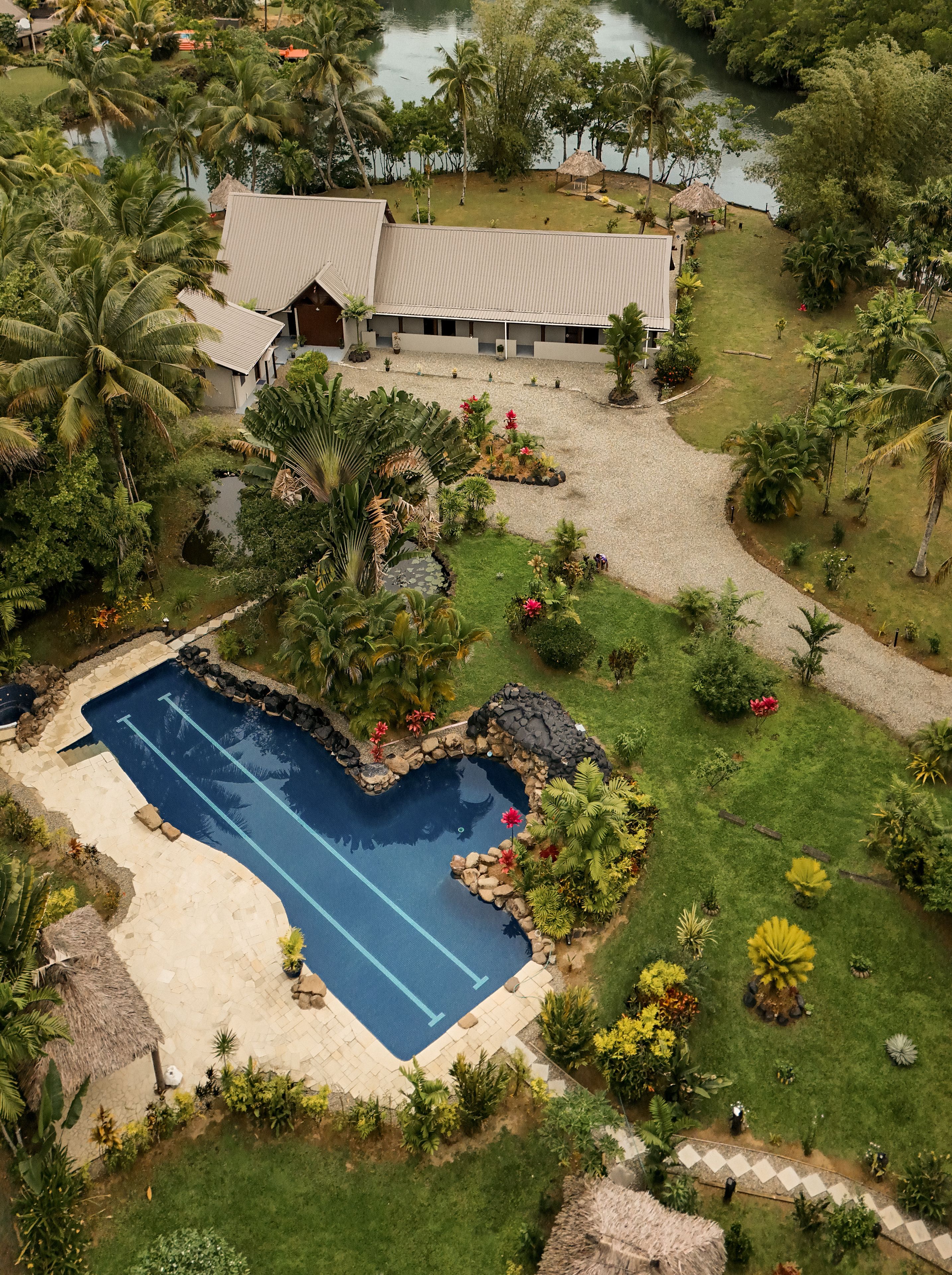 An aerial view of a resort with a swimming pool, lush greenery, and a large building with a thatched roof.