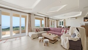 Un salon spacieux, doté d’un grand canapé, d’une table basse et offrant une vue sur l’extérieur grâce à de grandes portes-fenêtres coulissantes.