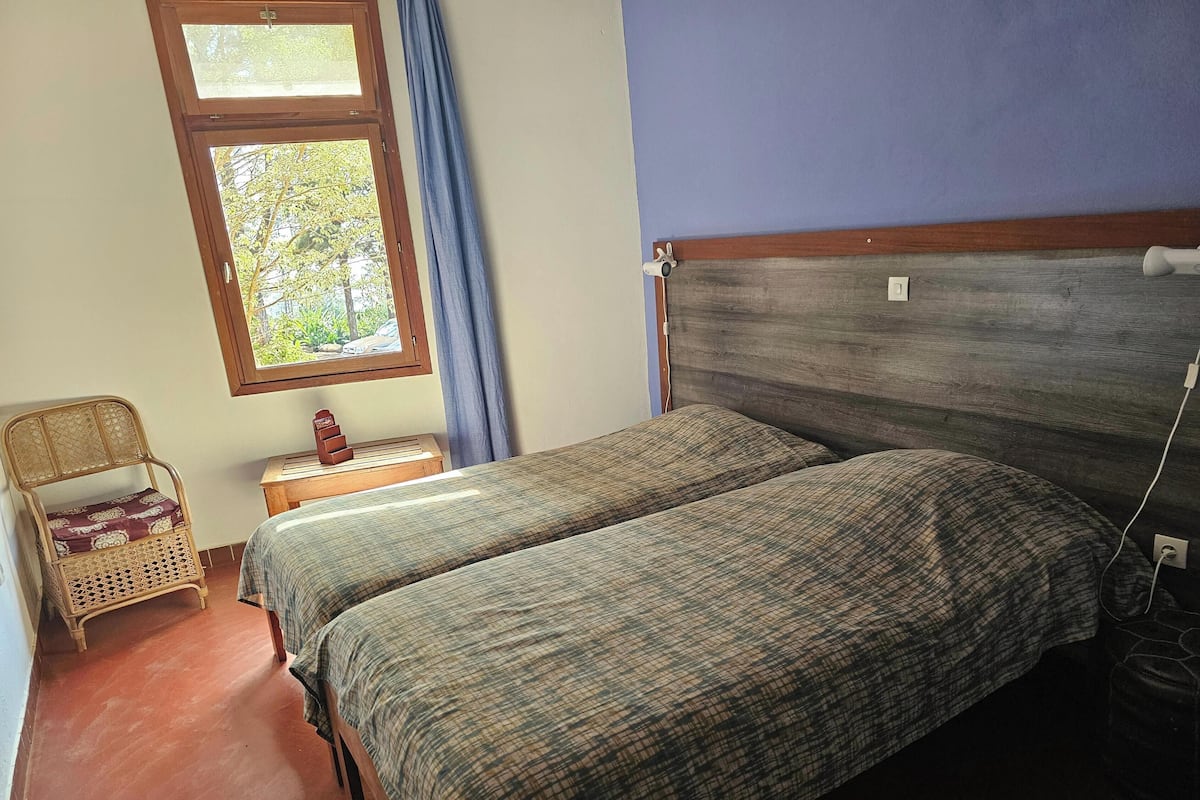 Un dormitorio con dos camas, cabecero de madera, ventana con vistas a árboles, una silla de mimbre y una mesita de noche con un reloj.