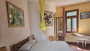 A bedroom with a bed, a desk, and two chairs. There are paintings on the walls and a window with curtains. - VILLA BOUTANGA (Bangoulap)