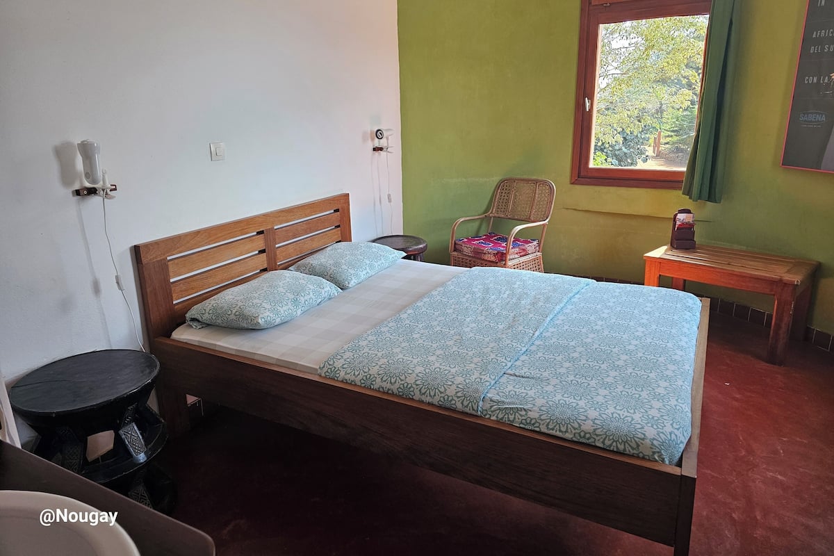 Una cama individual con cabecera de madera, una mesita de noche pequeña, una silla con un cojín estampado y una ventana con vista a árboles.