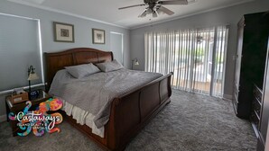 Un dormitorio con una cama grande, un ventilador de techo, una mesita de noche con lámpara y una ventana con persianas.