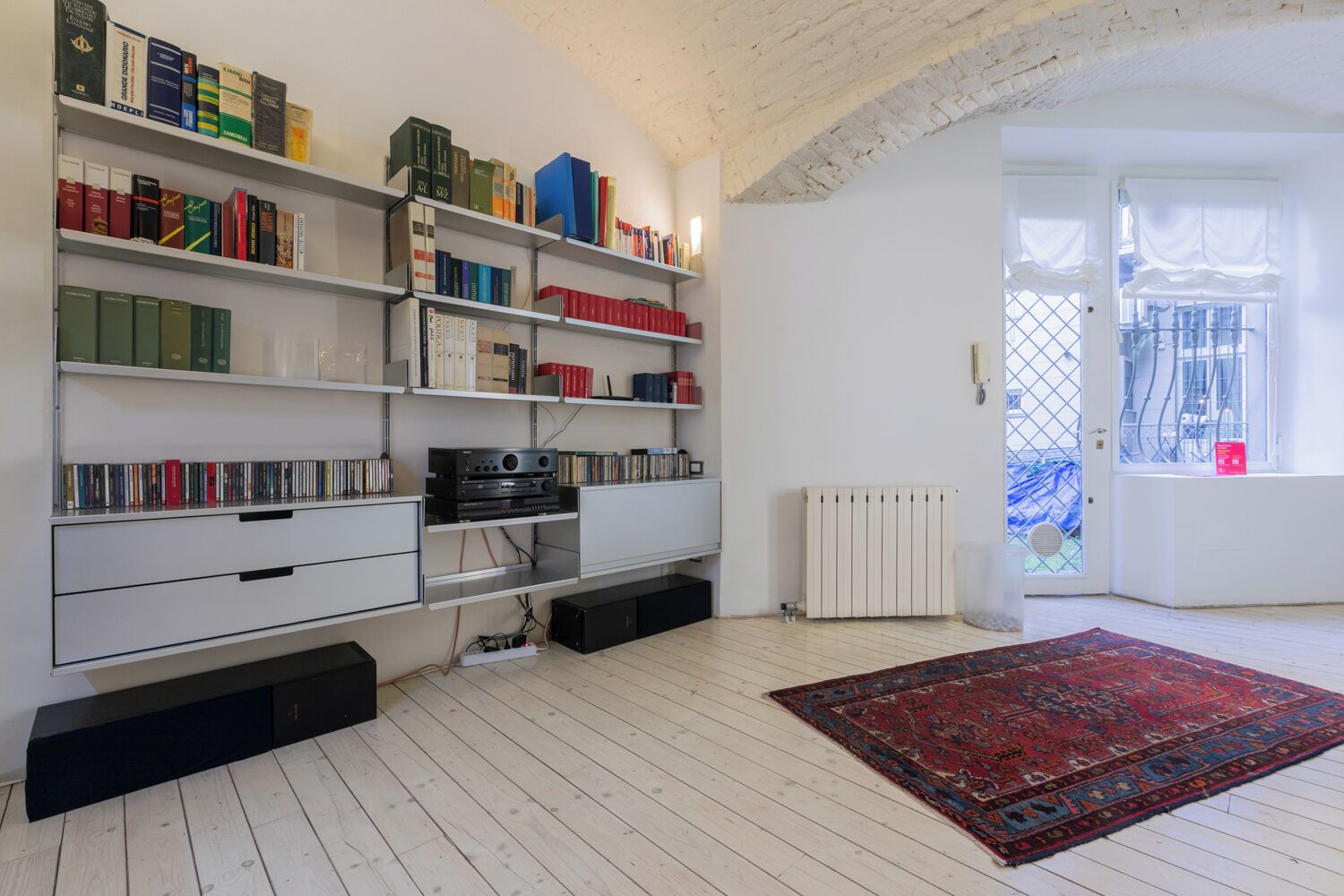 A room with white wooden flooring, a red patterned rug, and a bookshelf filled with books and DVDs.