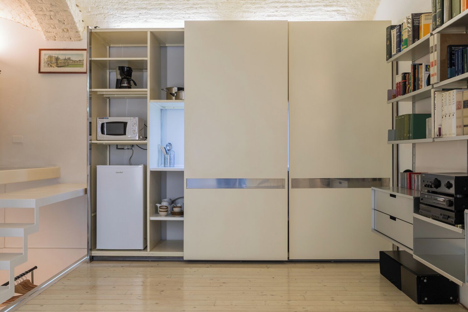 A compact kitchen with a microwave, a small refrigerator, and a bookshelf filled with books.