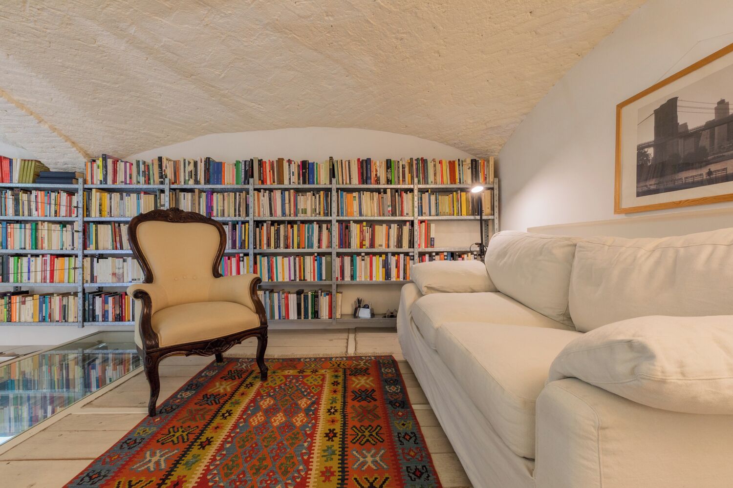 Un rincón acogedor para leer, con un sofá blanco, un sillón de madera y una estantería llena de libros.