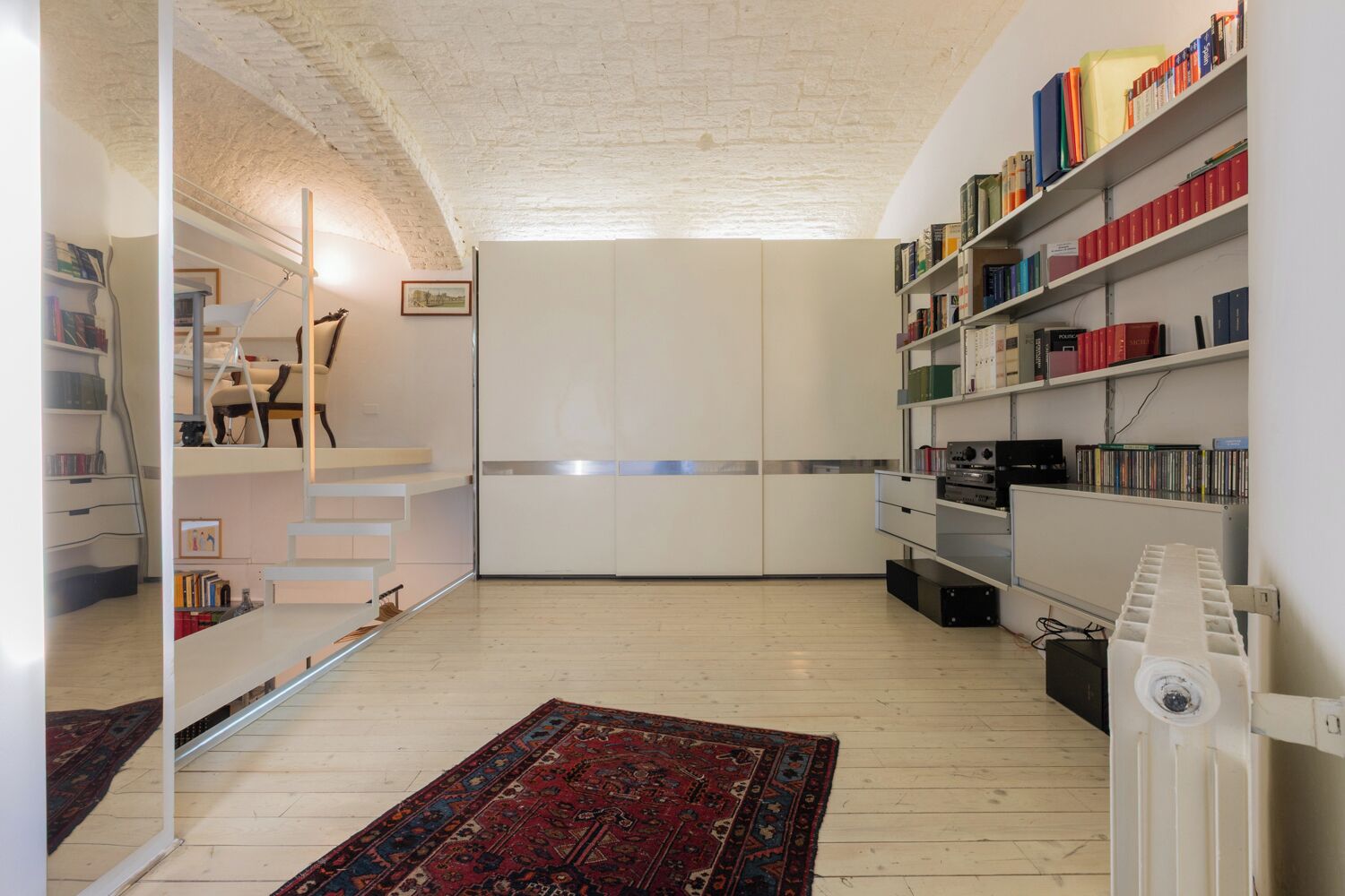 Una habitación amplia con techo alto, estantería empotrada, radiador y alfombra estampada.