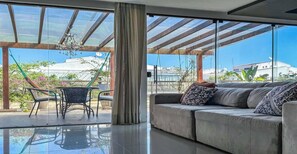 A modern living room with a large glass door leading to an outdoor area, a sofa with patterned cushions, and a dining table with chairs.