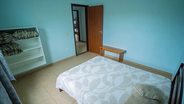 Quarto com cama, um banquinho, guarda-roupa e porta que dá para outro cômodo.