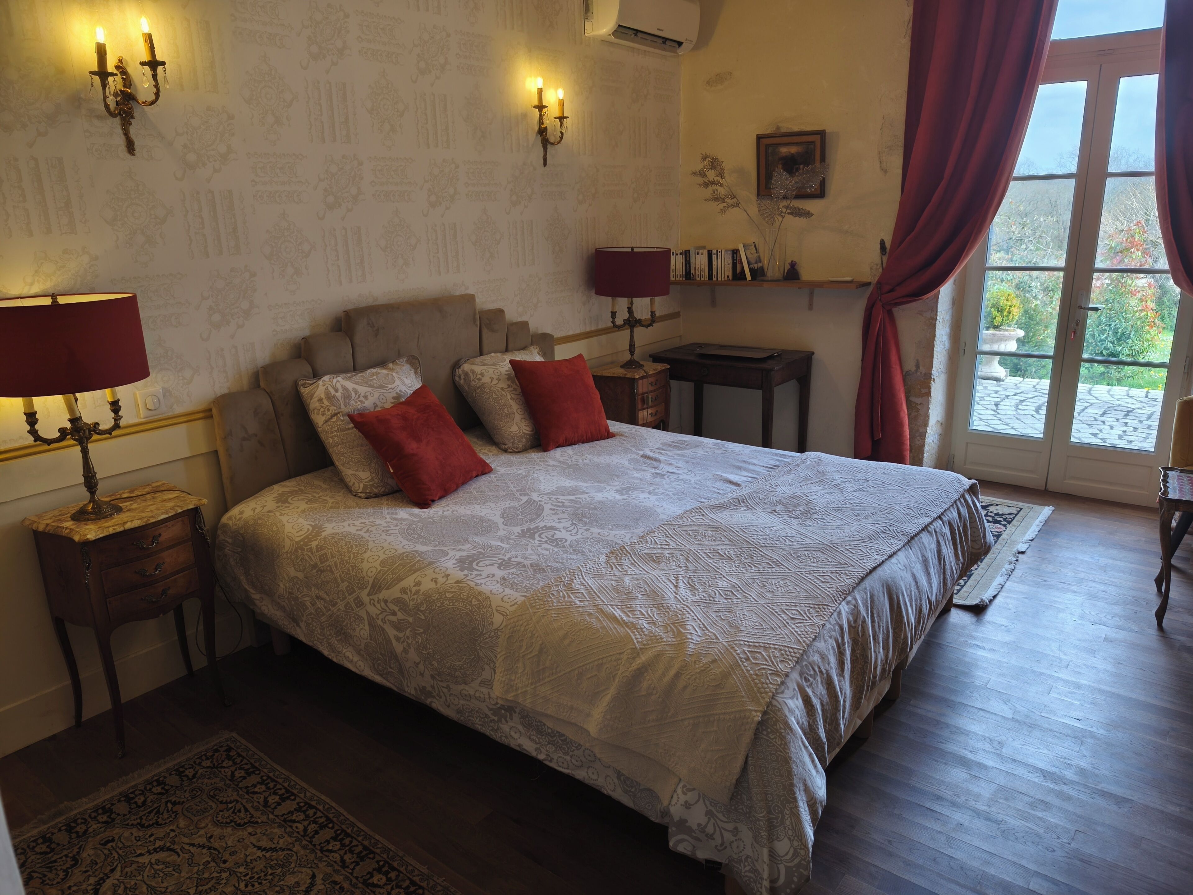 A bedroom with a large bed, two bedside tables, a nightstand, a lamp, a candelabra, a window with curtains, and a view of greenery outside.