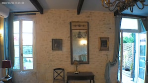 A bedroom with a bed, a chair, a table, and a mirror. There are two windows and a door leading to an outdoor area.