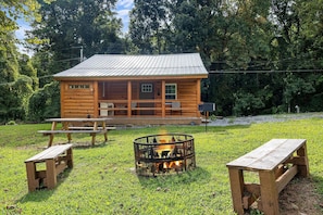 A wooden cabin with a metal roof, surrounded by a grassy area with picnic tables and a fire pit. - The Homestead 2 Bedroom Cabin (Cosby)