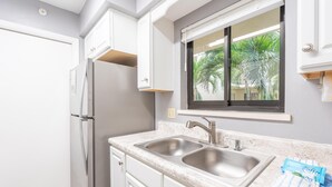 A kitchen with white cabinets, a double sink, and a window with a view of palm trees. - Santa Maria II #111 (Fort Myers Beach)