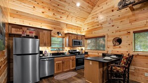A modern kitchen with wooden walls, stainless steel appliances, and a dining area with a table and chairs. - Cabin On A Hill 2 Bedroom Cabin (Cosby)