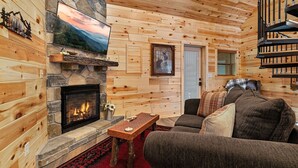 A cozy log cabin interior with a stone fireplace, wooden walls, a television mounted above it, and a comfortable seating area with a couch and pillows. - Cabin On A Hill 2 Bedroom Cabin (Cosby)